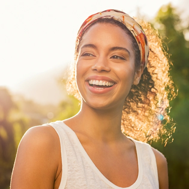 Young woman smiling outdoors in golden sunlight wearing white tank top and headband.