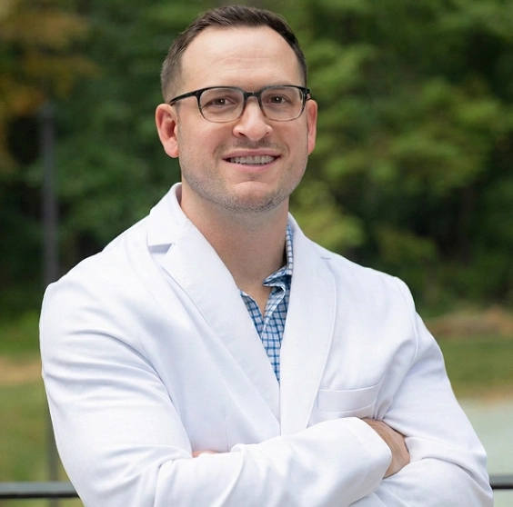 Male dental professional in white coat and glasses smiling outdoors with green background.